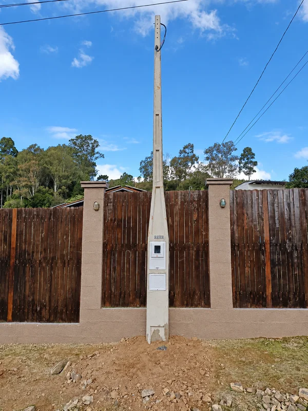 Poste de concreto com caixa de medição instalado diante de um portão de madeira.